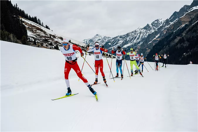 Eine Gruppe von Skisportlern fährt auf einer verschneiten Strecke in den Bergen. Im Hintergrund sind beeindruckende schneebedeckte Berge zu sehen.