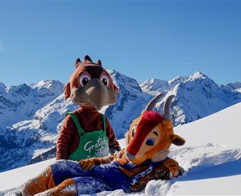 Zwei Maskottchen stehen im Schnee vor einer beeindruckenden Berglandschaft. Der Himmel ist klar und blau, während die schneebedeckten Berge im Hintergrund sichtbar sind.