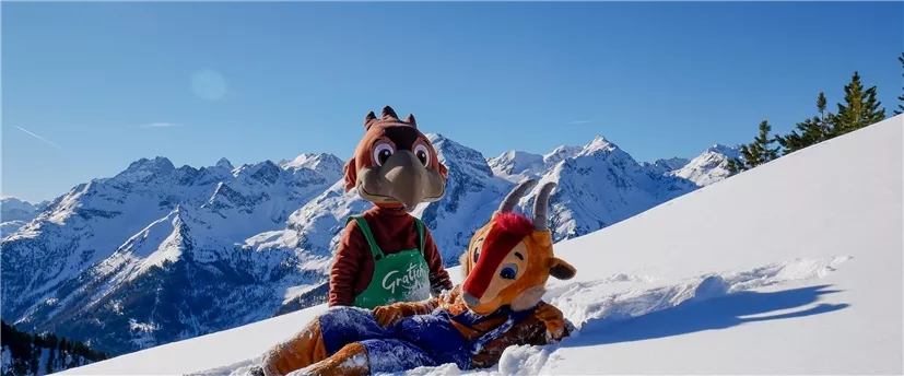 Zwei Maskottchen stehen im Schnee vor einer beeindruckenden Berglandschaft. Der Himmel ist klar und blau, während die schneebedeckten Berge im Hintergrund sichtbar sind.