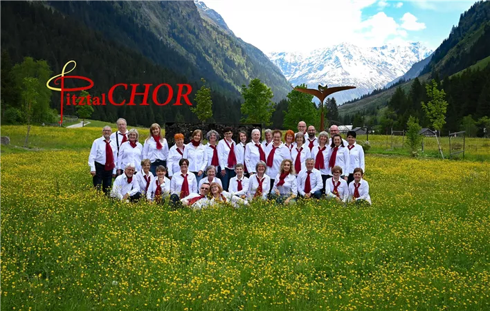 A group of singers stands in a blooming field in front of an impressive mountain landscape. All are wearing elegant clothing in white and red.