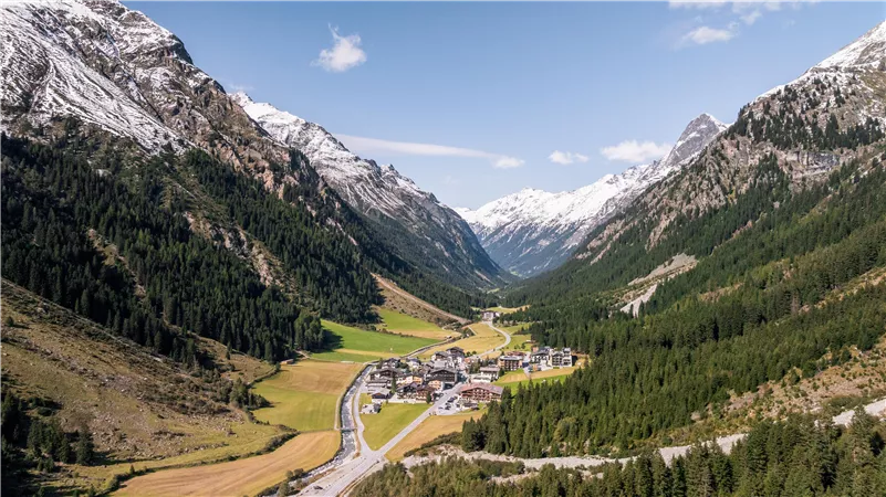 Eine malerische Berglandschaft mit schneebedeckten Gipfeln und einem kleinen Dorf im Tal. Rundherum sind grüne Wiesen und Wälder zu sehen.