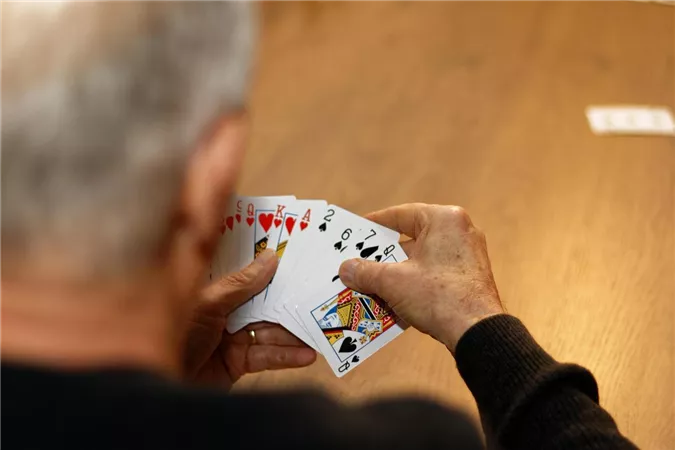 Eine Person hält eine Handkarten mit verschiedenen Spielkarten in den Händen. Der Tisch ist aus Holz und die Szene vermittelt eine entspannte Spielatmosphäre.