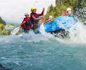 Zwei Radfahrer auf einem schmalen Pfad in einer grünen, hügeligen Landschaft mit Bergen im Hintergrund. Der Himmel ist blau mit einigen Wolken.