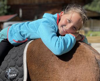 A smiling girl is lying relaxed on the back of a horse. In the background, wooden houses and a green environment are visible.