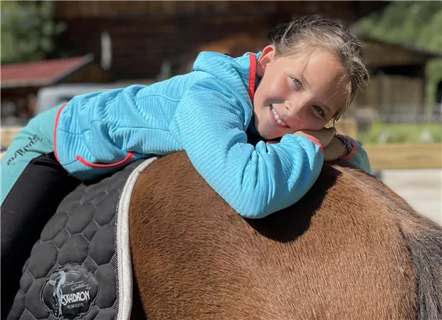 A smiling girl is lying relaxed on the back of a horse. In the background, wooden houses and a green environment are visible.