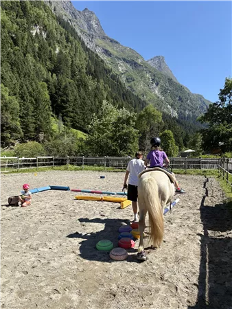 A riding arena in the midst of mountains, where a child is sitting on a horse. In the background, trees and a clear blue sky are visible.