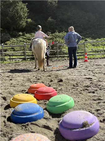 A pony with a rider stands on a training ground. In the foreground, colorful game discs are visible on the ground.