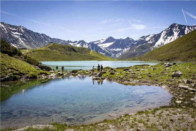 Ein malerischer Bergsee mit glitzertem Wasser und schneebedeckten Gipfeln im Hintergrund. Grünes Gras und Felsen umgeben das Ufer, während Menschen am Wasser stehen.