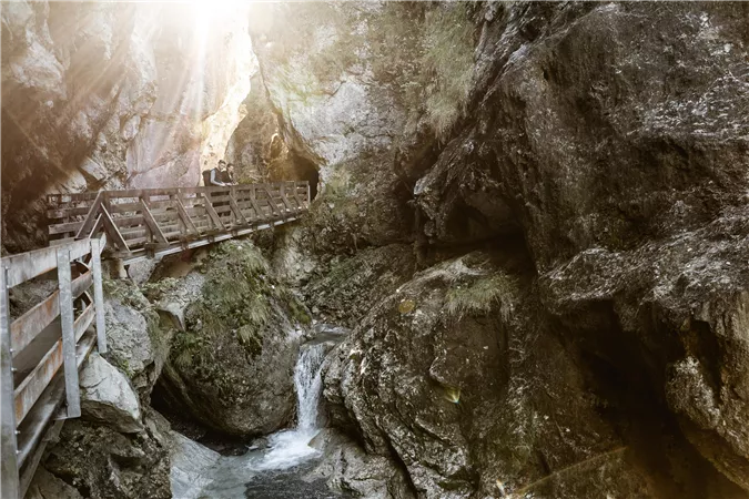 A charming house built against a rock, surrounded by green trees. The sky is cloudy and the surroundings seem calm and rural.