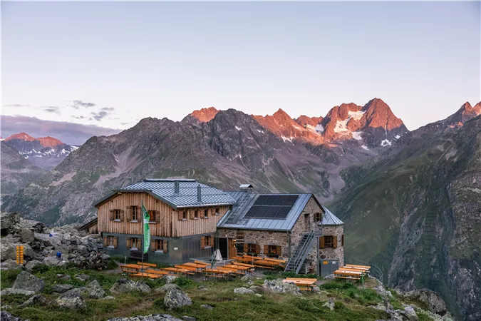 Eine Berghütte umgeben von majestätischen Bergen und einer schönen Landschaft. Die sanften Farben des Sonnenuntergangs verleihen der Szene einen warmen Glanz.