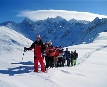 A group of hikers stands in the snow and walks uphill. In the background, majestic mountains can be seen under a clear blue sky.
