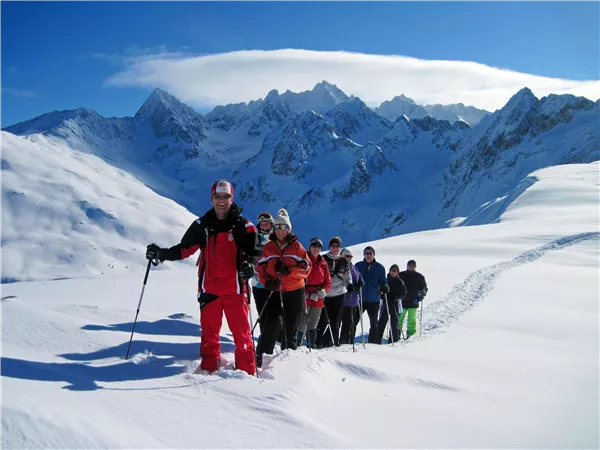 A group of hikers stands in the snow and walks uphill. In the background, majestic mountains can be seen under a clear blue sky.