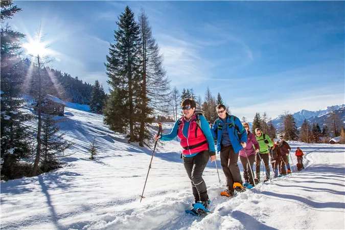 Eine Gruppe von Menschen wandert mit Schneeschuhen durch eine verschneite Landschaft. Die Sonne scheint klar am Himmel und umgibt die Szene mit einem schönen Licht.
