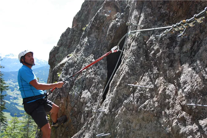 A man is climbing a steep rock face with safety hooks. In the background, green forests and mountains can be seen.