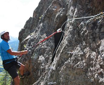 A man is climbing a steep rock face with safety hooks. In the background, green forests and mountains can be seen.
