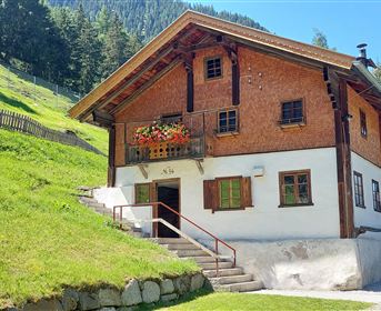 Ein hübsches Haus mit Holzfassade und Balkon, umgeben von grünem Gras und Bäumen. Im Hintergrund sind Berge sichtbar.