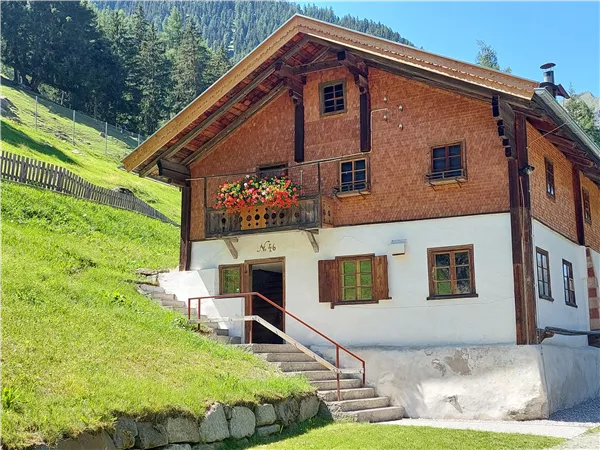 Ein hübsches Haus mit Holzfassade und Balkon, umgeben von grünem Gras und Bäumen. Im Hintergrund sind Berge sichtbar.