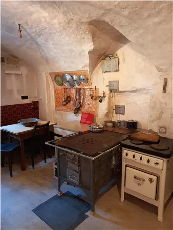 Eine rustikale Küche mit einer alten Kochstelle und einem Tisch. Die Wände sind aus Stein und es hängen verschiedene Küchenutensilien an der Wand.