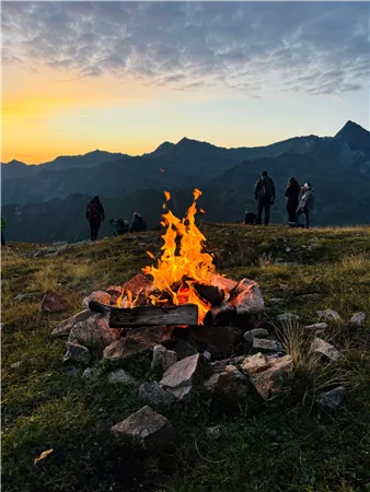 Ein Lagerfeuer brennt auf einer Wiese mit Bergen im Hintergrund. Im Sonnenuntergang sind einige Personen zu sehen, die die Aussicht genießen.