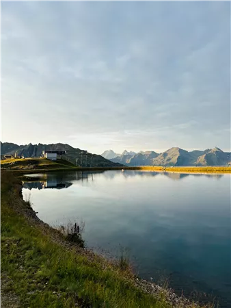 Ein ruhiger See umgeben von Bergen und Gras. Der Himmel ist leicht bewölkt und spiegelt sich im Wasser.