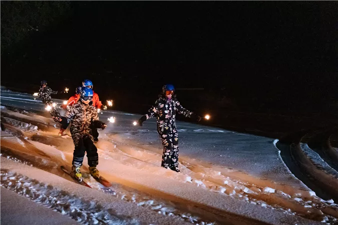 Eine Gruppe von Skifahrern fährt nachts mit Fackeln durch den Schnee. Die Teilnehmer tragen bunte Skikleidung und Helme.