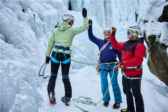 Drei Kletterer feiern gemeinsam am Fuße eines gefrorenen Wasserfalls. Sie tragen Kletterausrüstung und erheben die Hände zum High-Five.