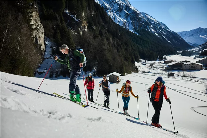 Eine Gruppe von Skitourengehern wandert durch verschneite Berge. Die Sonne scheint und die Landschaft wirkt malerisch.
