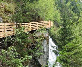 A wooden path runs along a steep slope with lush greenery. A waterfall flows down beside the path.