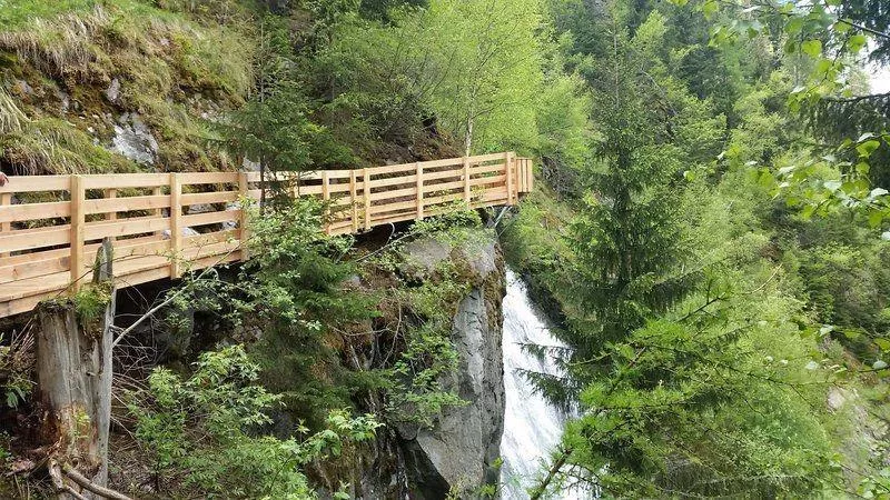 Ein Holzweg führt entlang eines steilen Abhangs mit üppigem Grün. Neben dem Weg fließt ein Wasserfall herab.