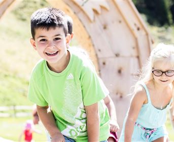 Zwei Kinder spielen draußen auf einem Spielplatz. Im Hintergrund sind grüne Wiesen und Bäume sichtbar.