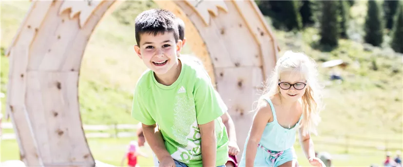 Zwei Kinder spielen draußen auf einem Spielplatz. Im Hintergrund sind grüne Wiesen und Bäume sichtbar.