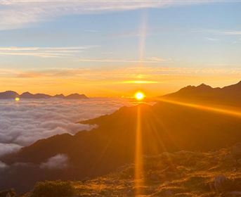 Ein atemberaubender Sonnenaufgang über den Bergen, mit einer goldenen Sonne, die über den Wolken aufgeht. Die Landschaft ist von sanften Hügeln und einem klaren Himmel umgeben.