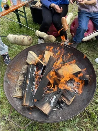 A cozy campfire with flickering flames and fresh wood. People are roasting sausages over the fire.
