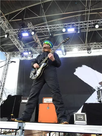 Ein Musiker spielt Gitarre auf einer Bühne. Er trägt eine schwarze Kleidung und Sonnenbrille.