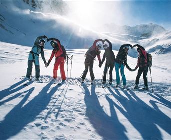 A group of six people stands in the snow, forming a heart with their hands. Snow-covered mountains and a clear blue sky can be seen in the background.