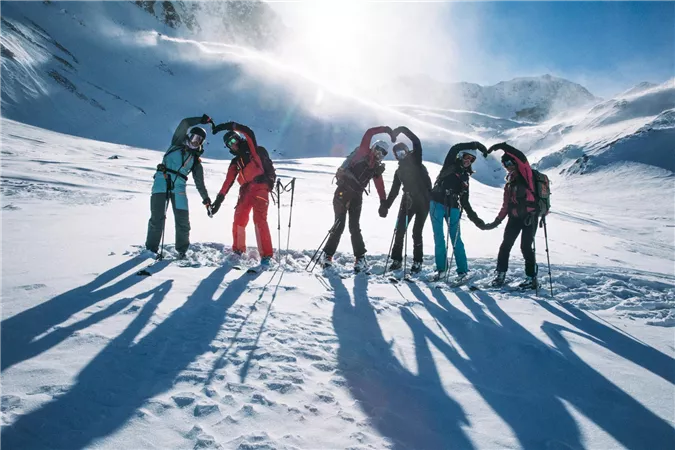 Eine Gruppe von sechs Personen steht im Schnee und bildet mit ihren Händen ein Herz. Im Hintergrund sind schneebedeckte Berge und ein klarer blauer Himmel zu sehen.
