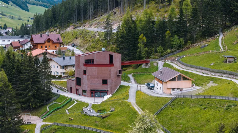 Eine moderne, rechteckige Gebäude mit rotem Mauerwerk umgeben von grünen Wiesen und einer Waldlandschaft. Im Hintergrund sind mehrere traditionelle Häuser zu sehen.