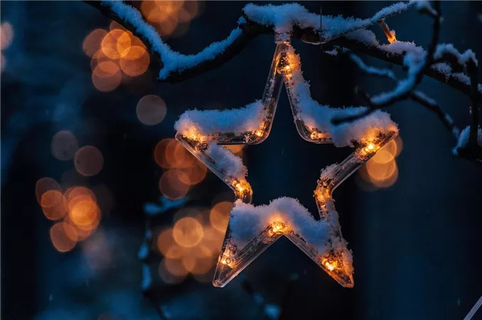 Ein leuchtender Weihnachtsstern hängt an einem Ast, der mit Schnee bedeckt ist. Im Hintergrund sind unscharfe Lichter zu sehen, die eine festliche Stimmung erzeugen.
