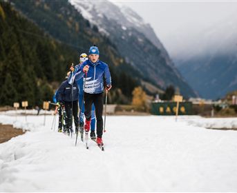 A group of people is cross-country skiing through a snow-covered landscape. In the background, there are mountains and partially forested areas visible.