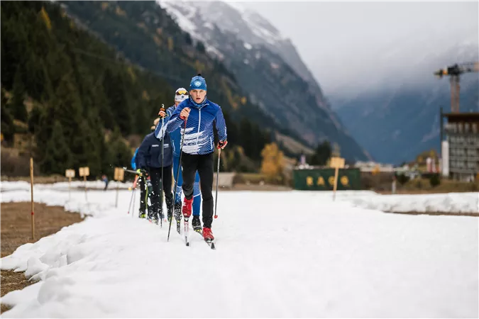 A group of people is cross-country skiing through a snow-covered landscape. In the background, there are mountains and partially forested areas visible.