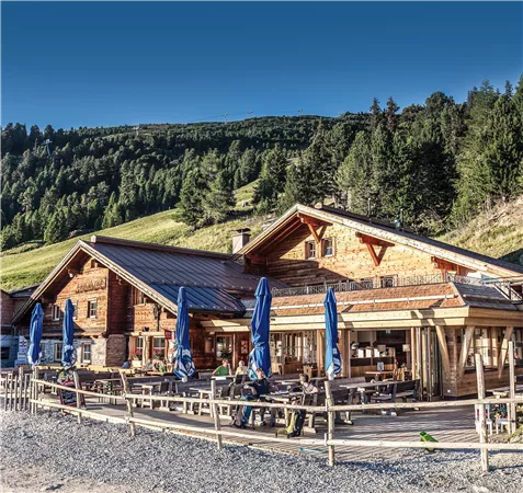 A picturesque mountain house with a wooden facade and terrace. Surrounded by green forests and a clear sky.