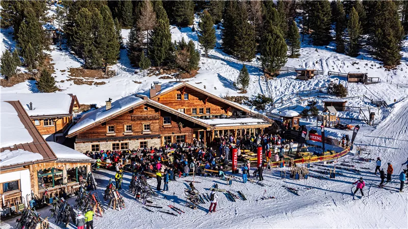 Eine belebte Skihütte in den Bergen mit vielen Menschen. Umgeben von Schnee und grünen Bäumen, bietet die Hütte einen gemütlichen Ort zum Entspannen.