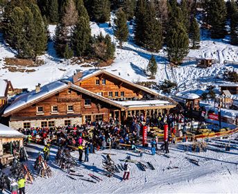 Ein geschäftiges Alpinhütte mit vielen Menschen auf einer verschneiten Wiese. Im Hintergrund sind hohe Bäume und eine Skispur zu sehen.