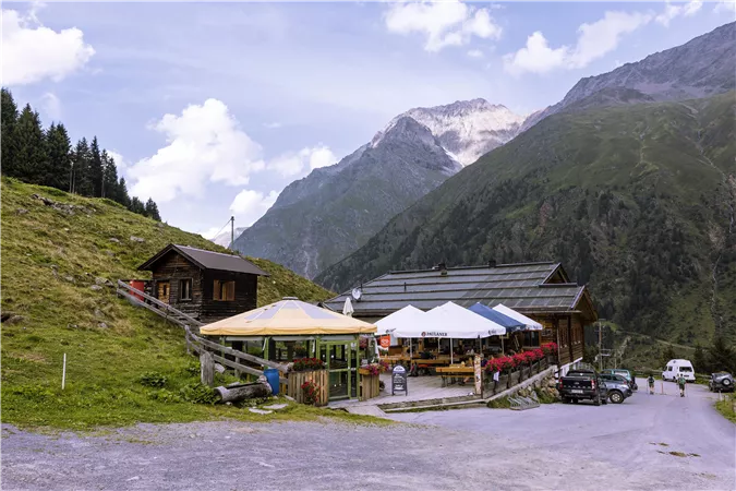 Eine malerische Berghütte mit Terrassen im Freien und umgeben von hohen Bergen. Der Himmel ist klar und die Natur ist grün und einladend.