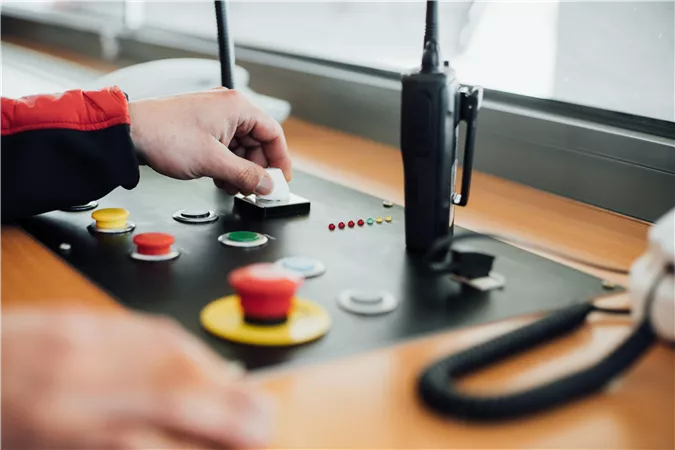 Eine Hand bedient ein Kontrollpanel mit verschiedenen Knöpfen in verschiedenen Farben. Im Hintergrund ist ein Funkgerät sichtbar.