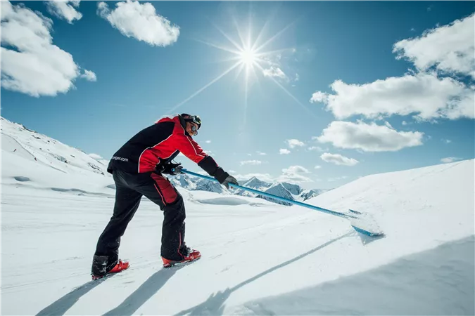 Eine Person hebt Schnee mit einer Schaufel auf einer sonnigen Gebirgslandschaft. Der Himmel ist blau und mit einigen Wolken bedeckt.