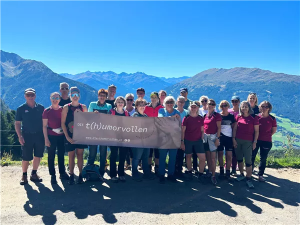 Eine Gruppe von Menschen steht im Freien und hält ein Banner. Im Hintergrund sind Berge und ein klarer, blauer Himmel zu sehen.