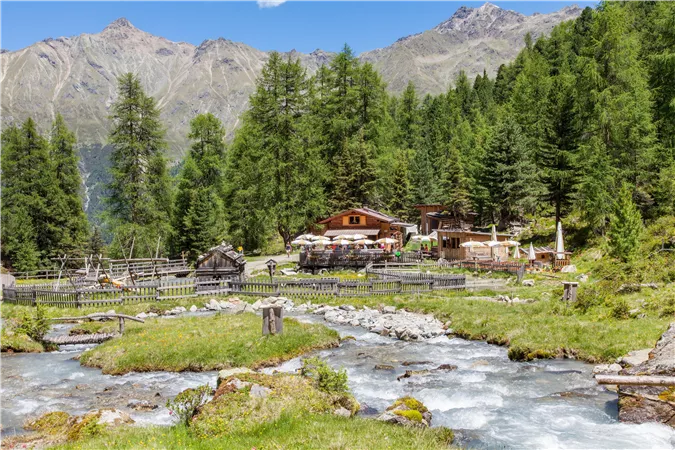 A picturesque high mountain landscape with green meadows and dense forests. In the background, cozy wooden houses and a clear river can be seen.
