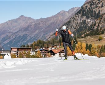 Eine Person, die auf einem schneebedeckten Terrain Ski läuft, umgeben von majestätischen Bergen. Im Hintergrund sind einige alpine Hütten sichtbar.
