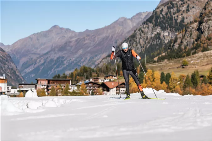 Eine Person, die auf einem schneebedeckten Terrain Ski läuft, umgeben von majestätischen Bergen. Im Hintergrund sind einige alpine Hütten sichtbar.